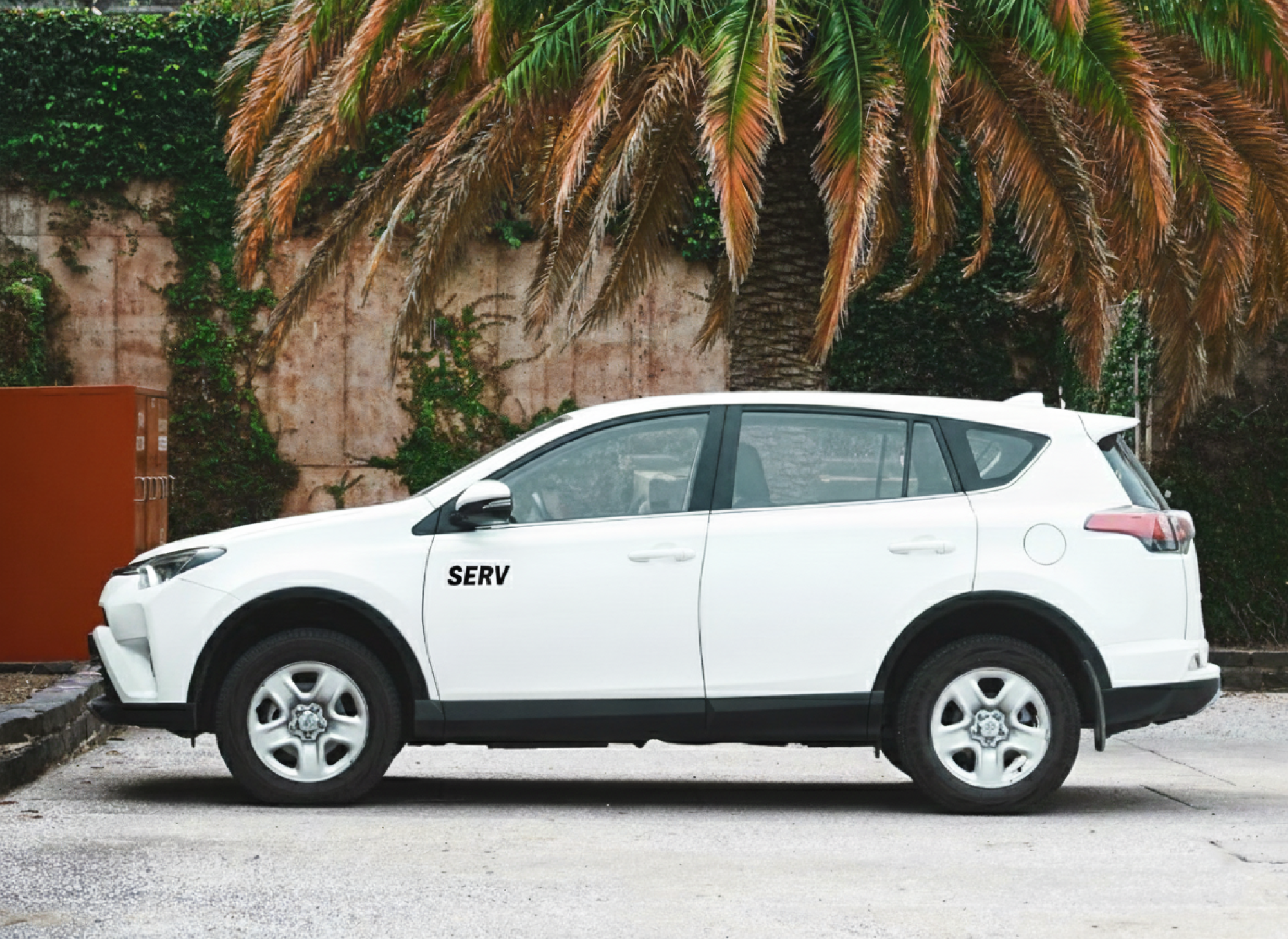 A white SUV parked by a concrete lot with a palm-fringed wall and ivy, labeled &ldquo;SERV,&rdquo; under a sunny, tropical backdrop.