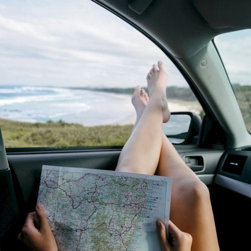 A person relaxing in a car with legs up on the dashboard, holding a map, ocean view outside the window, sunny day.