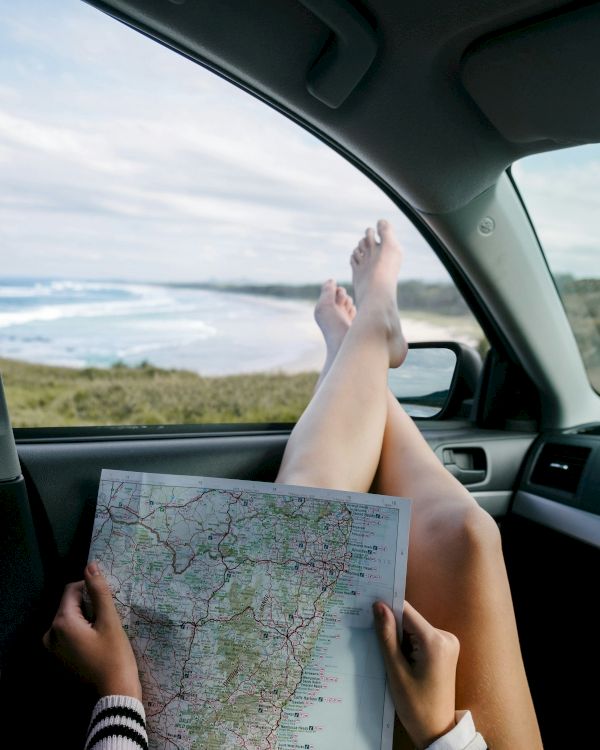 A person relaxing in a car with legs up on the dashboard, holding a map, ocean view outside the window, sunny day.