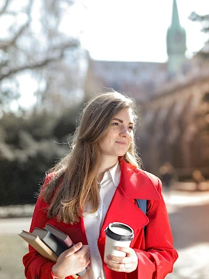 A young woman in a red coat holds books and a coffee cup outdoors on a sunny campus path.