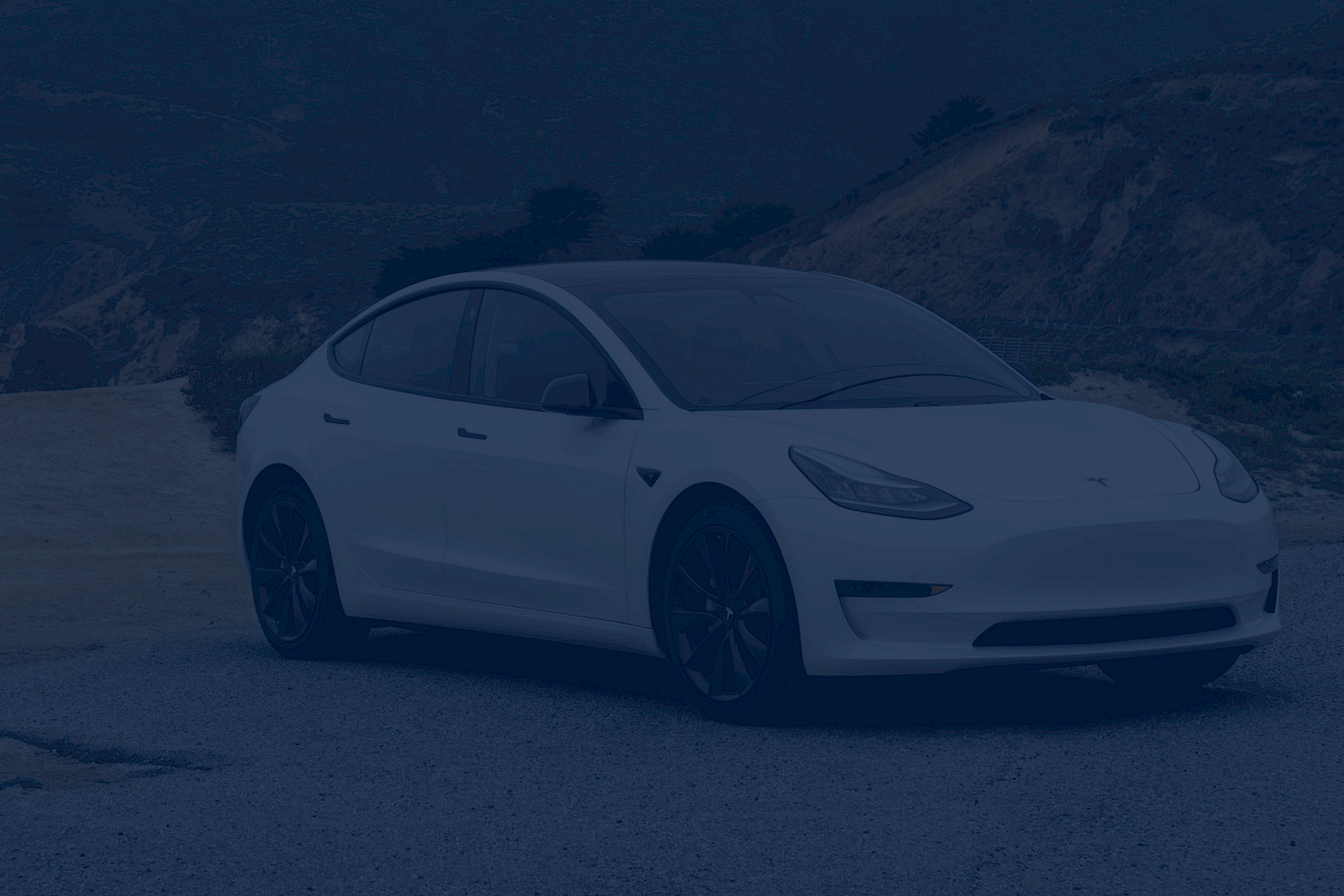 A white Tesla Model Y parked on a scenic cliffside road with coastal hills in the background.