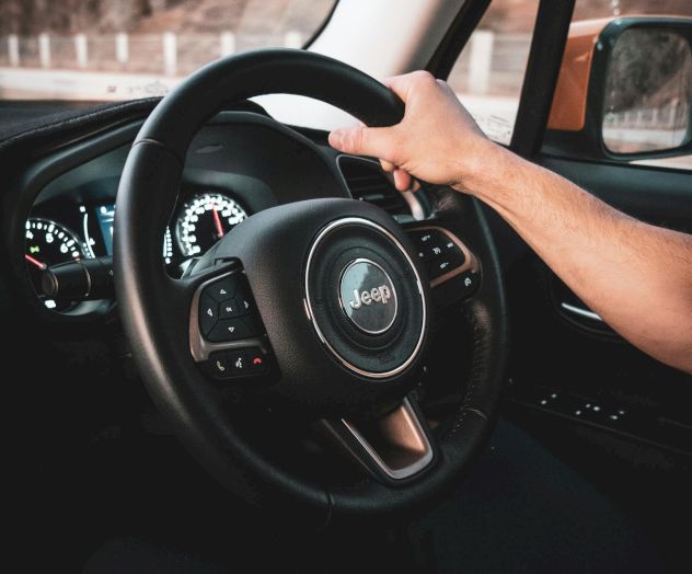 A close-up of a car steering wheel, with hands gripping it; dashboard details and a circular horn emblem in the center.