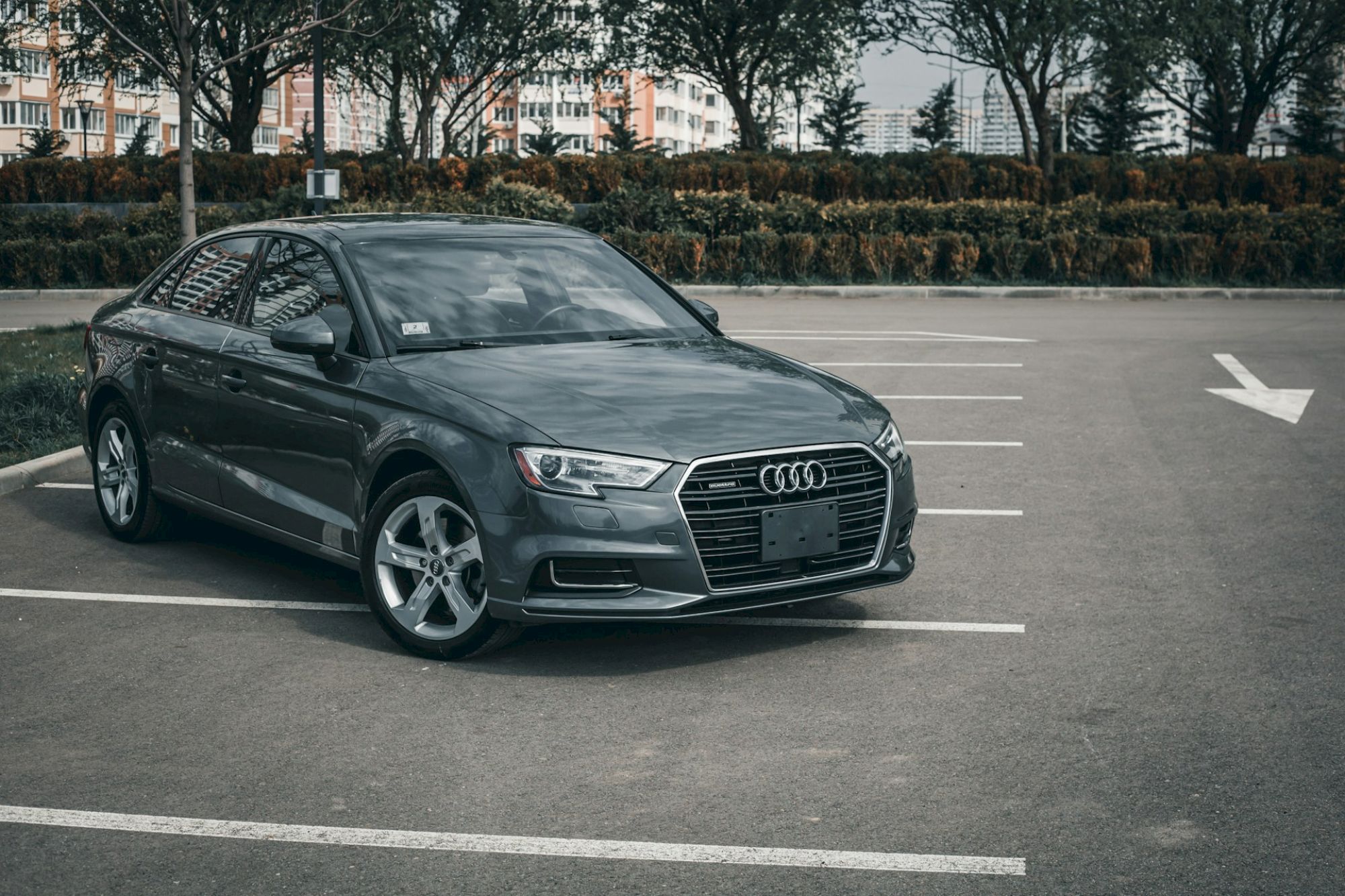 A dark gray Audi sedan parked in a mostly empty parking lot, with white lines and a right-turn arrow in the background.